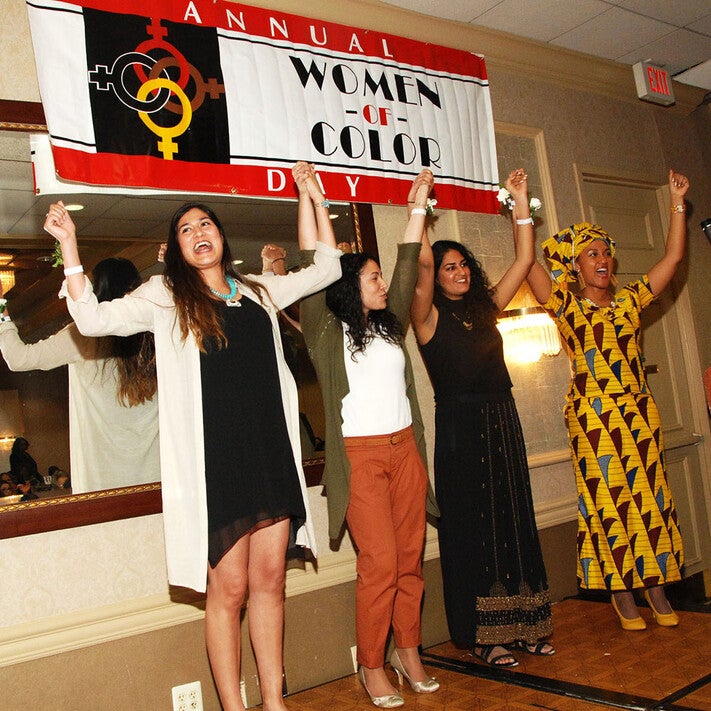 smiling women of color with arms raised in front of Annual Women of Color Day banner